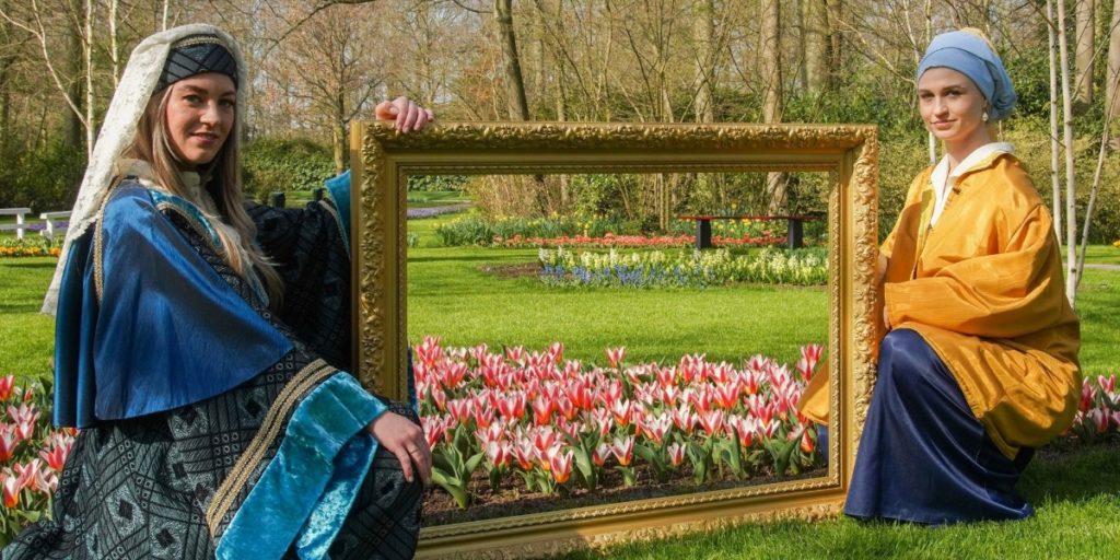 Two young ladies, dressed up in traditional Dutch clothing from the 1700 and 1800. They are sitting on the grass holding a frame of a painting. There is no painting inside but behind it you will see the grass and the tulips growing in Keukenhof gardens.